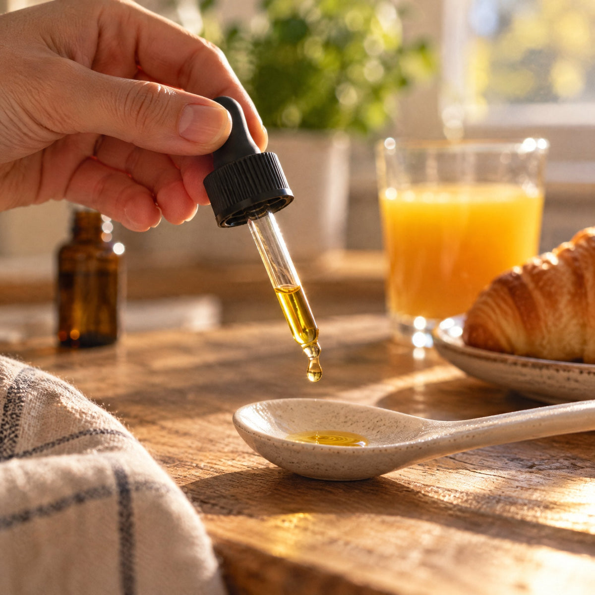 Hand haelt eine Pipette, ein Tropfen faellt auf einen Keramikloeffel, Fruehstueckstisch im Goldlicht, die Flasche steht unscharf im Hintergrund