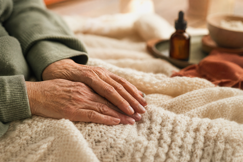 Entspannte Hände einer älteren Person auf einer cremefarbenen Strickdecke, im Hintergrund unscharf eine Braunglas-Pipettenflasche