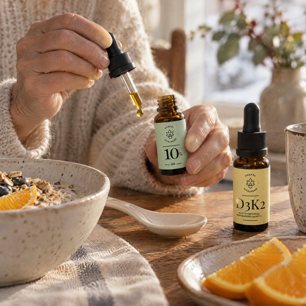 Hände geben einen Tropfen CBD-Öl 10 % am Frühstückstisch mit Porridge und Orangen, daneben das Vitamin D3+K2